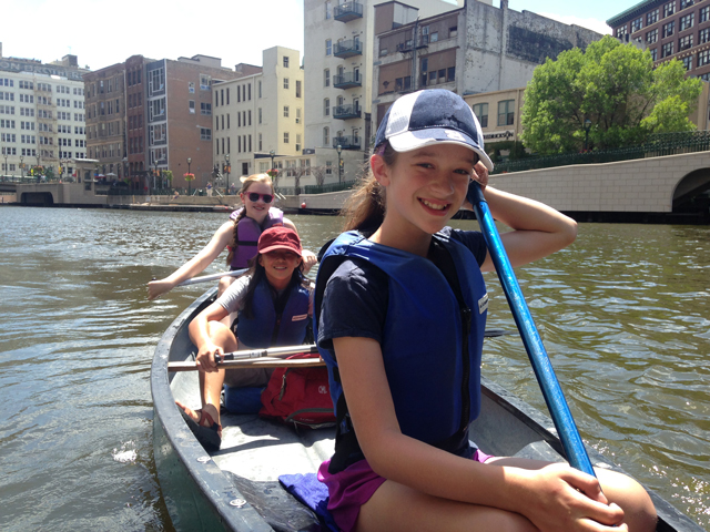 Girl in the front seat of a canoe, holds a paddle. Behind her are two other girls, one in the middle and one paddling in the rear seat.