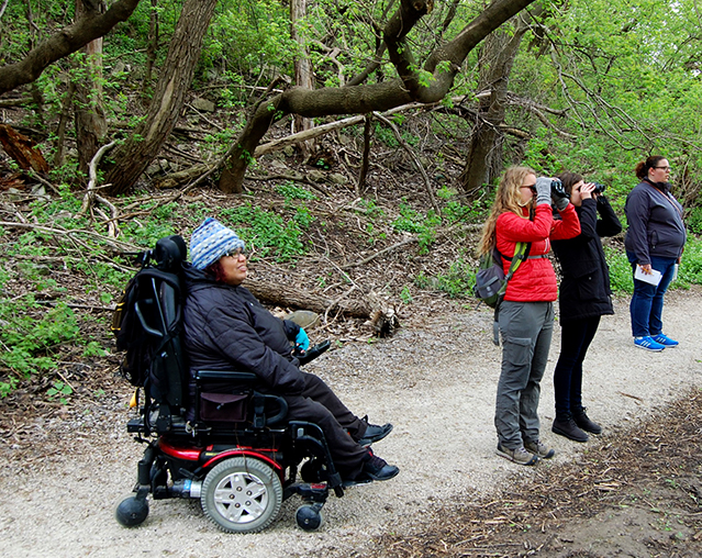 A woman in a power chair, two women looking through binoculars all on a pressed limestone path