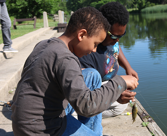 African American boy is sitting along the lagoon, smiling at the very first fish he's ever caught.