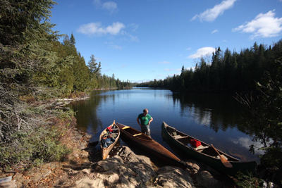 Ken standing in the water at the edge of a lake. He's surrounded by tall pines and is standing among the three canoes used on the trip.